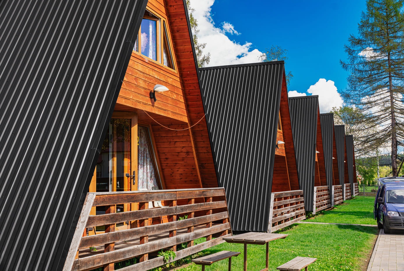 Szereg drewnianych domków z czarnym, trapezowym dachem na tle błękitnego nieba i zieleni. Widoczny fragment parkingu i niebieski samochód dostawczy.