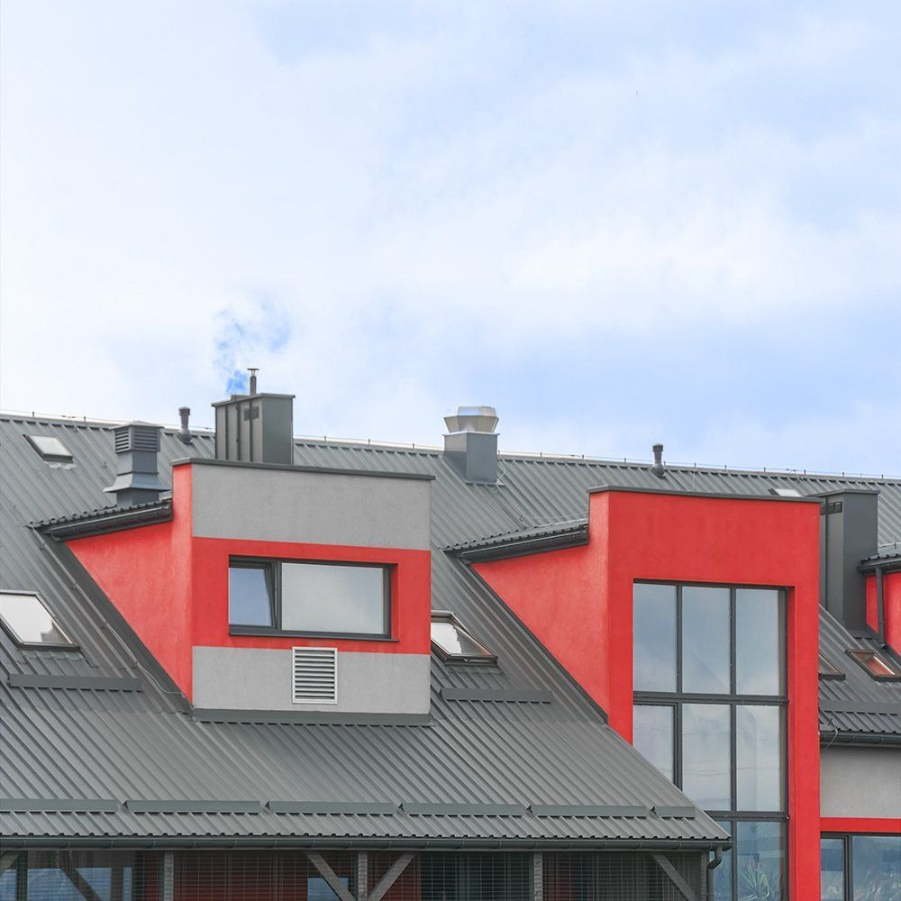 Szary dach z blachy trapezowej z czerwonymi nadbudówkami okiennymi i szarymi kominami na tle błękitnego nieba z lekkimi chmurami. Z komina unosi się dym.