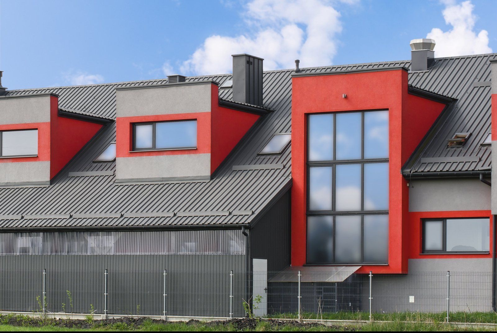 Szary dach z blachy trapezowej na budynku z czerwonymi i szarymi elementami architektonicznymi, widok z poziomu gruntu, błękitne niebo.