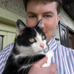 Psycholog Oleśnica Jakub Krasucki Prywatny Gabinet Psychologiczny Metanoja Poradnictwo Terapia Diagnoza osobowości - Mężczyzna w koszuli w paski trzyma czarno-białego kota na tle ściany z pnączami i ozdobnych drzwi z wyciętym sercem.