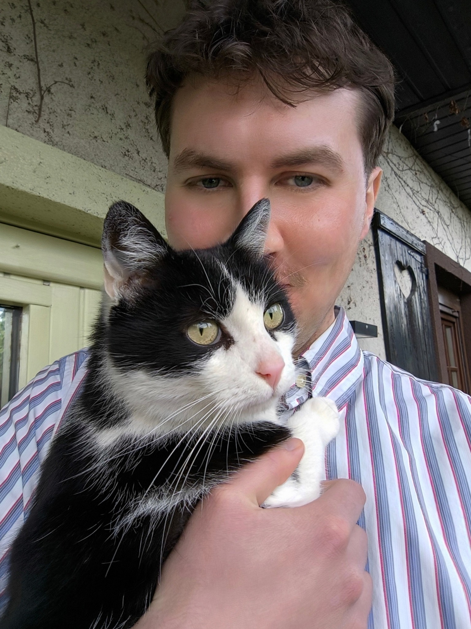 Mężczyzna w koszuli w paski trzyma czarno-białego kota na tle ściany z pnączami i ozdobnych drzwi z wyciętym sercem.