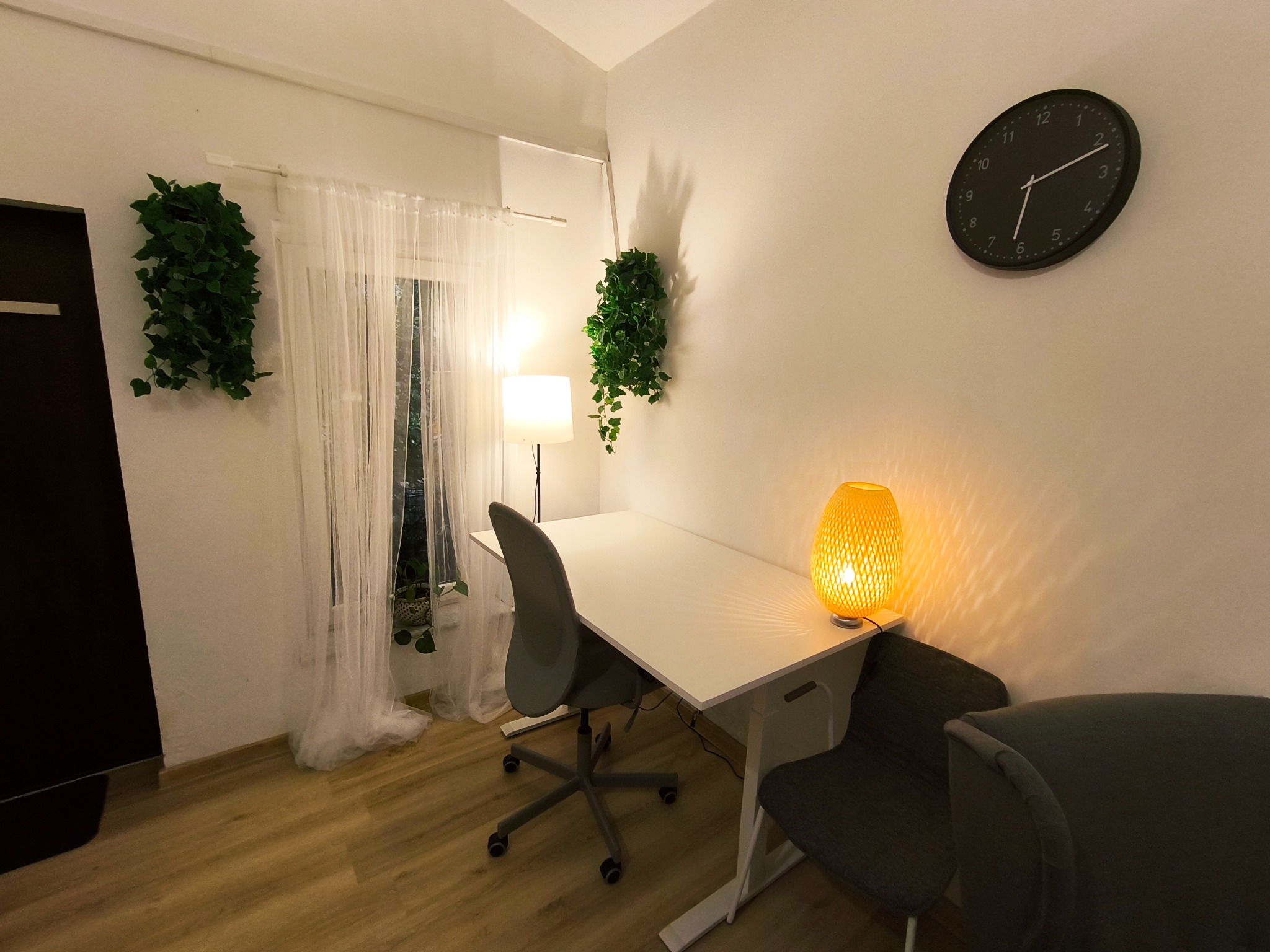 Jasny gabinet psychologiczny z biurkiem, krzesłami, lampą i zegarem na ścianie. Dwa pnącza i firanka przy oknie. Przytulne, spokojne wnętrze.