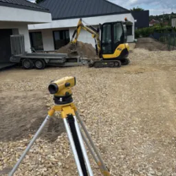 Żółty niwelator optyczny na statywie, w tle minikoparka Yanmar na żwirowym terenie budowy, dom z szarą dachówką i przyczepa. Błękitne niebo z chmurami.