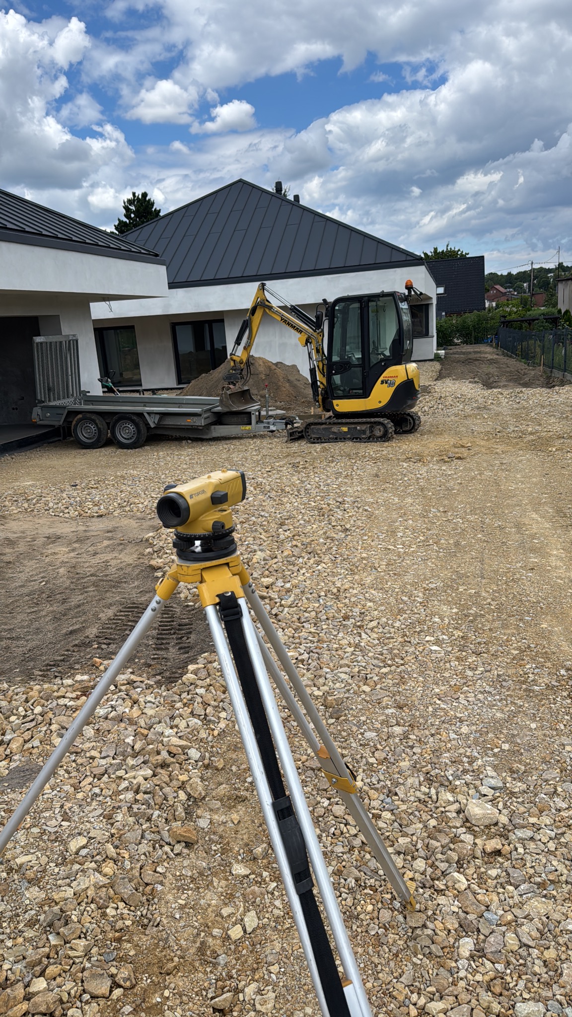 Żółty niwelator optyczny na statywie, w tle minikoparka Yanmar na żwirowym terenie budowy, dom z szarą dachówką i przyczepa. Błękitne niebo z chmurami.
