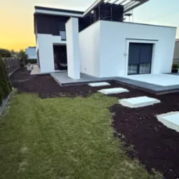 Nowoczesny dom z białymi ścianami i czarnym dachem, widok z ogrodu z częściowo ułożonymi płytami chodnikowymi i przygotowanym terenem pod trawnik o zachodzie słońca.