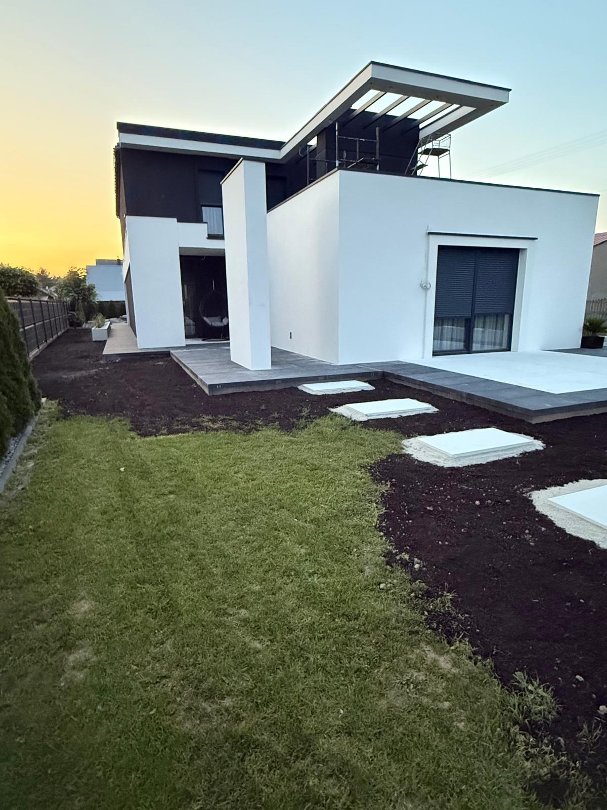 Nowoczesny dom z białymi ścianami i czarnym dachem, widok z ogrodu z częściowo ułożonymi płytami chodnikowymi i przygotowanym terenem pod trawnik o zachodzie słońca.