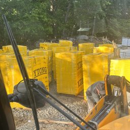 Piotr Hornicki - Widok z kabiny koparki na plac budowy z ułożonymi paletami bloczków Ytong, przygotowanymi do wznoszenia murów. W tle zalesiony teren.