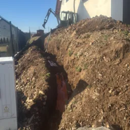 Wykopany wąski rów w ziemi z pomarańczową folią, obok domu. Na górze widać koparkę. Wykop pod instalacje.