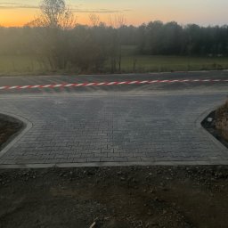 Piotr Hornicki - Ułożona szara kostka brukowa na rozwidleniu drogi, zabezpieczona taśmą ostrzegawczą; w tle drzewa i niebo o zachodzie słońca. Widoczne nierówności terenu po bokach.