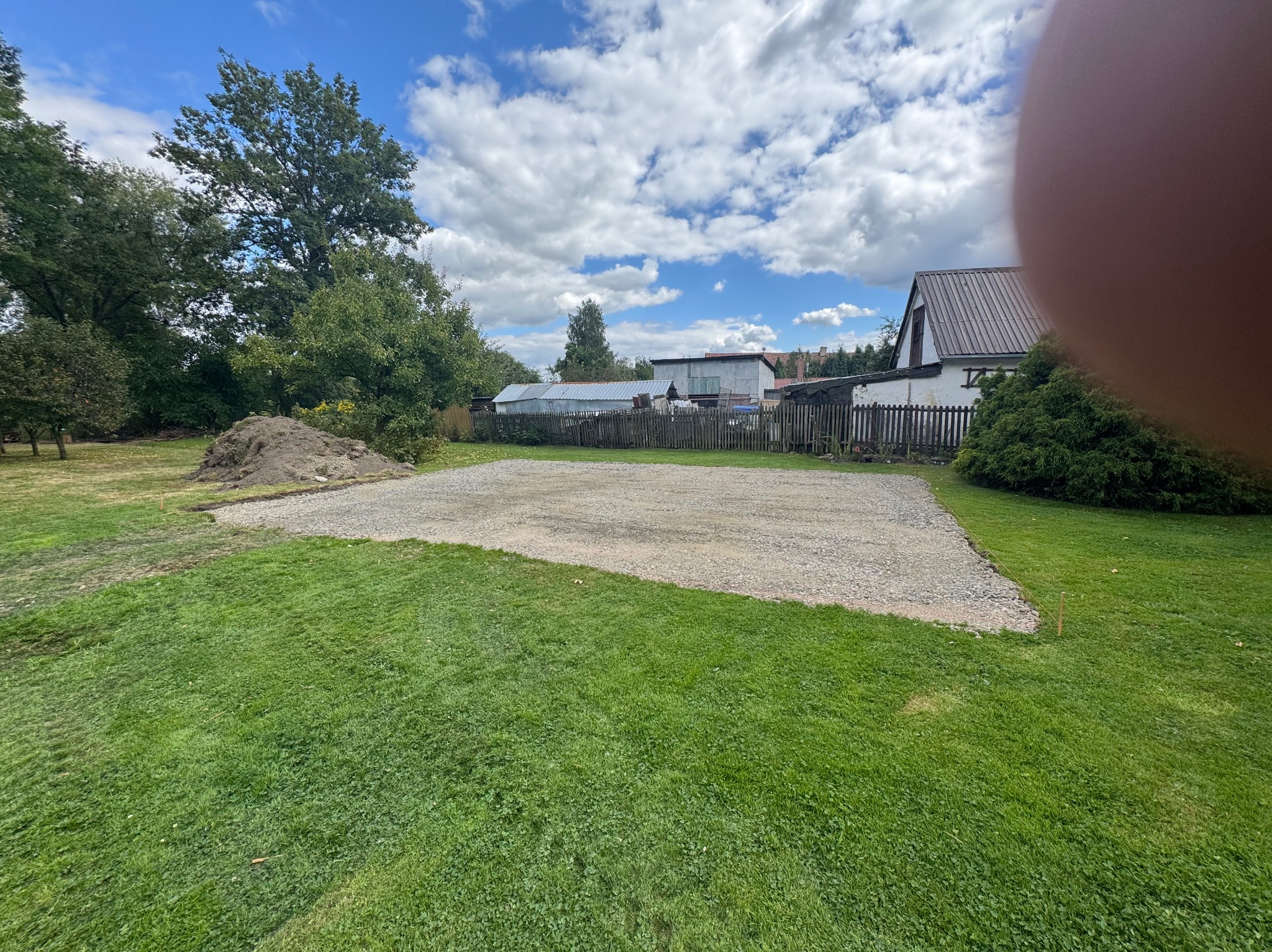Utwardzony teren żwirem na posesji, widoczne drzewa i budynki w tle, błękitne niebo z chmurami. Przygotowany plac pod przyszłą budowę lub parking.