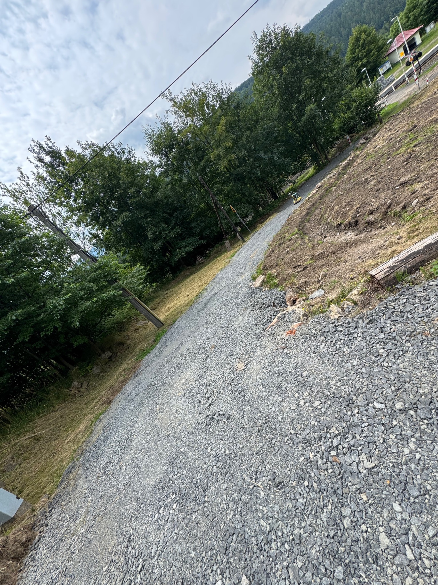 Utwardzona, żwirowa droga wzdłuż zbocza, z widoczną roślinnością i elementami infrastruktury kolejowej w tle. Prace ziemne w toku, z częściowo przygotowanym terenem.