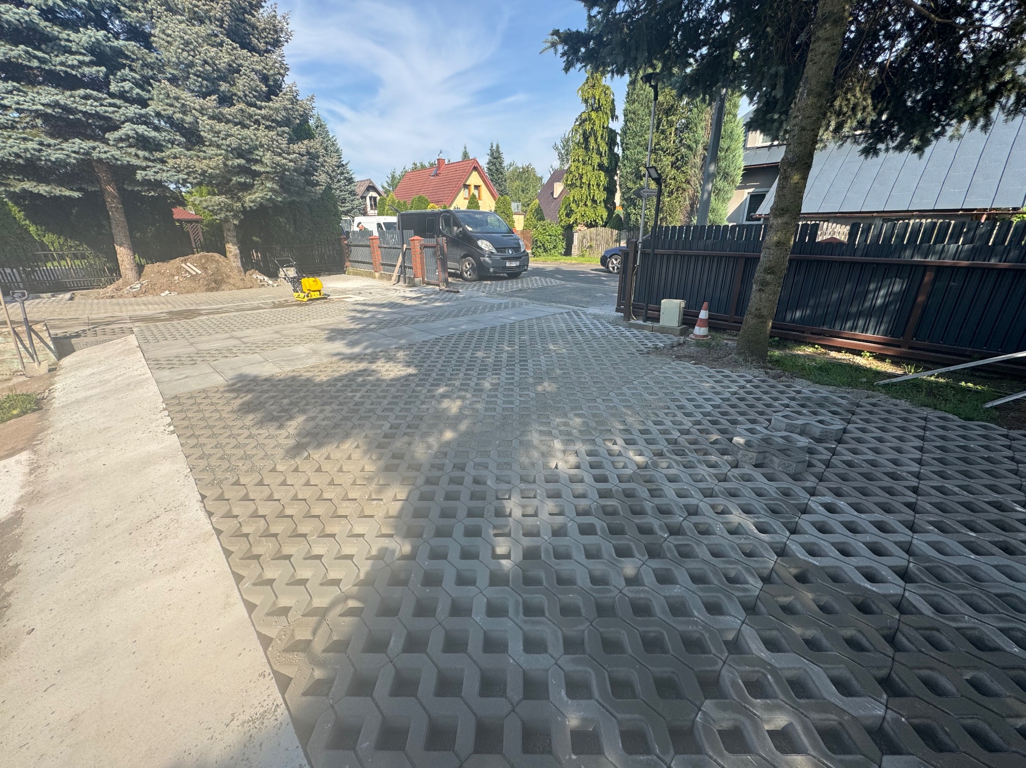 Ułożona, szara ażurowa kostka brukowa na podjeździe, z widocznym ubijakiem w tle. Częściowo widoczny samochód i ogrodzenie. Słoneczny dzień.