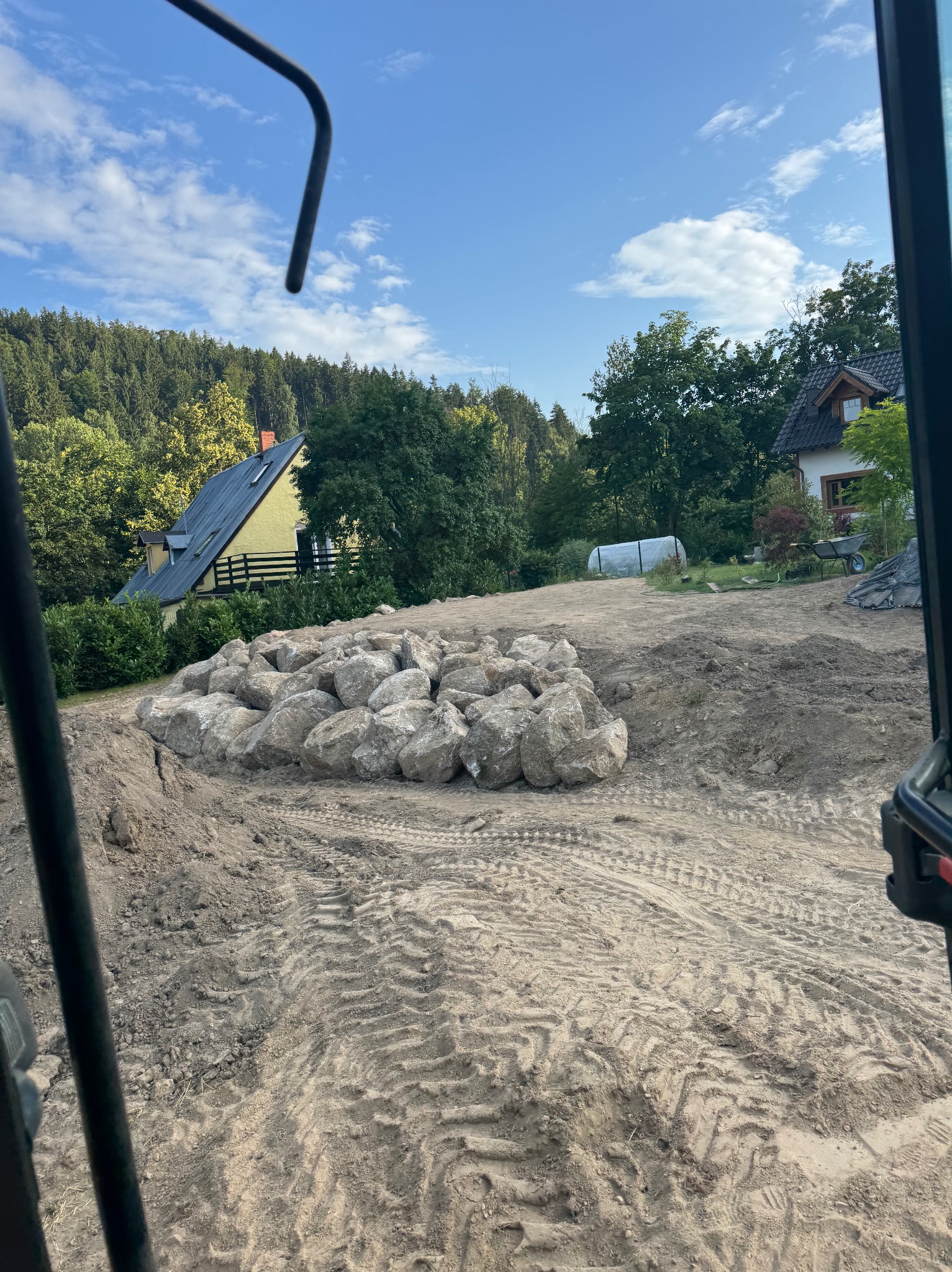 Przygotowany teren pod budowę z pryzmą kamieni, widok z kabiny maszyny budowlanej. W tle domy i zieleń.