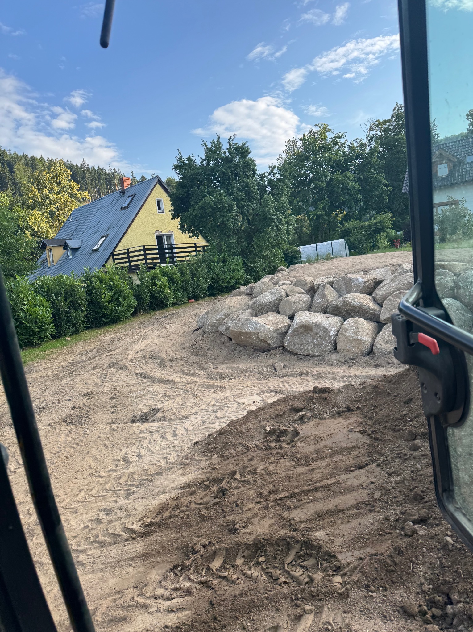 Widok z kabiny maszyny budowlanej na teren z usypanymi kamieniami, w tle żółty dom z szarym dachem i zieleń. Ślady opon na ziemi.