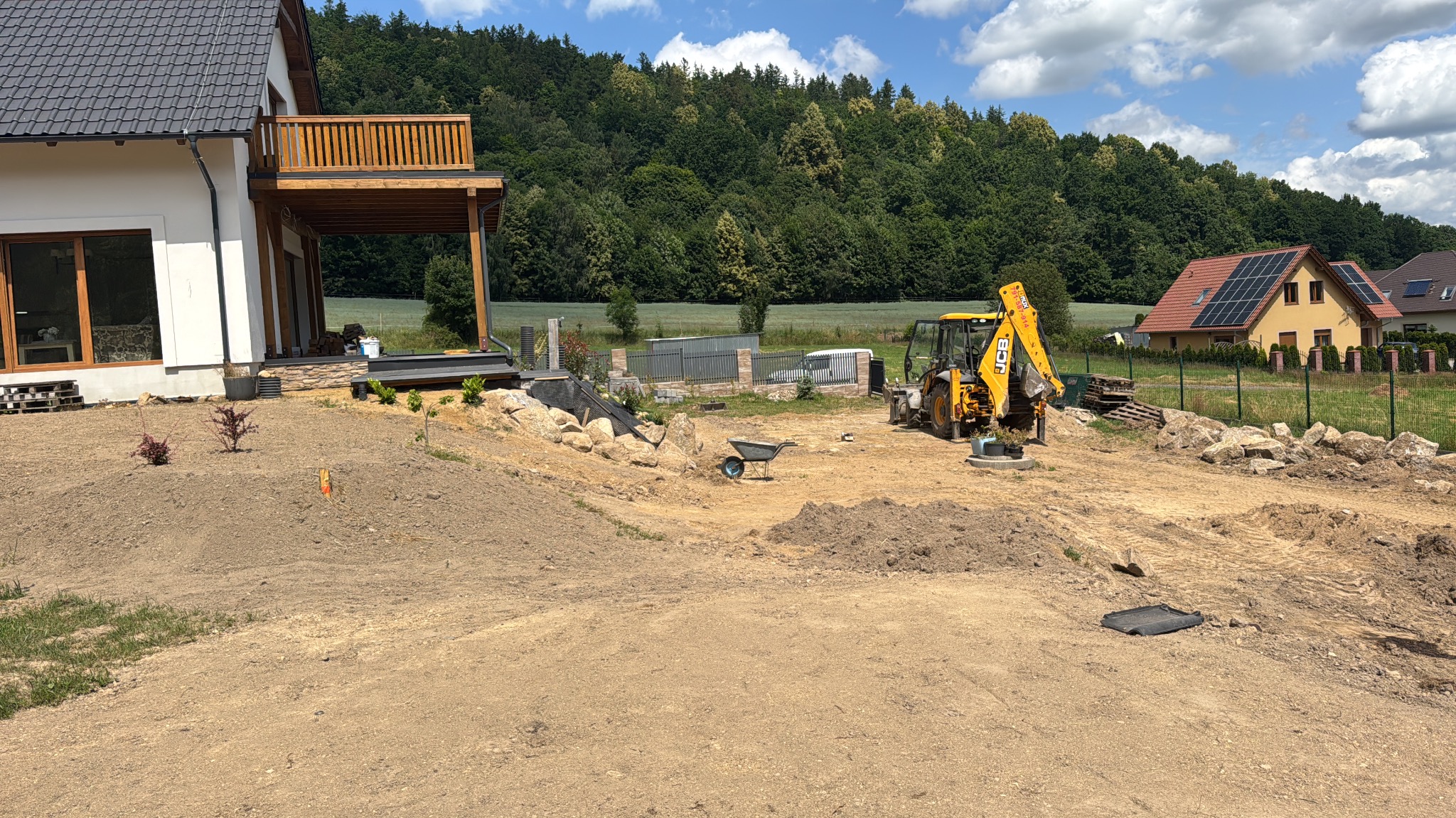 Żółta koparko-ładowarka JCB na działce w trakcie prac ziemnych, widoczny dom z drewnianym balkonem, ukształtowany teren z kamieniami. Słoneczny dzień, zielone drzewa w tle.