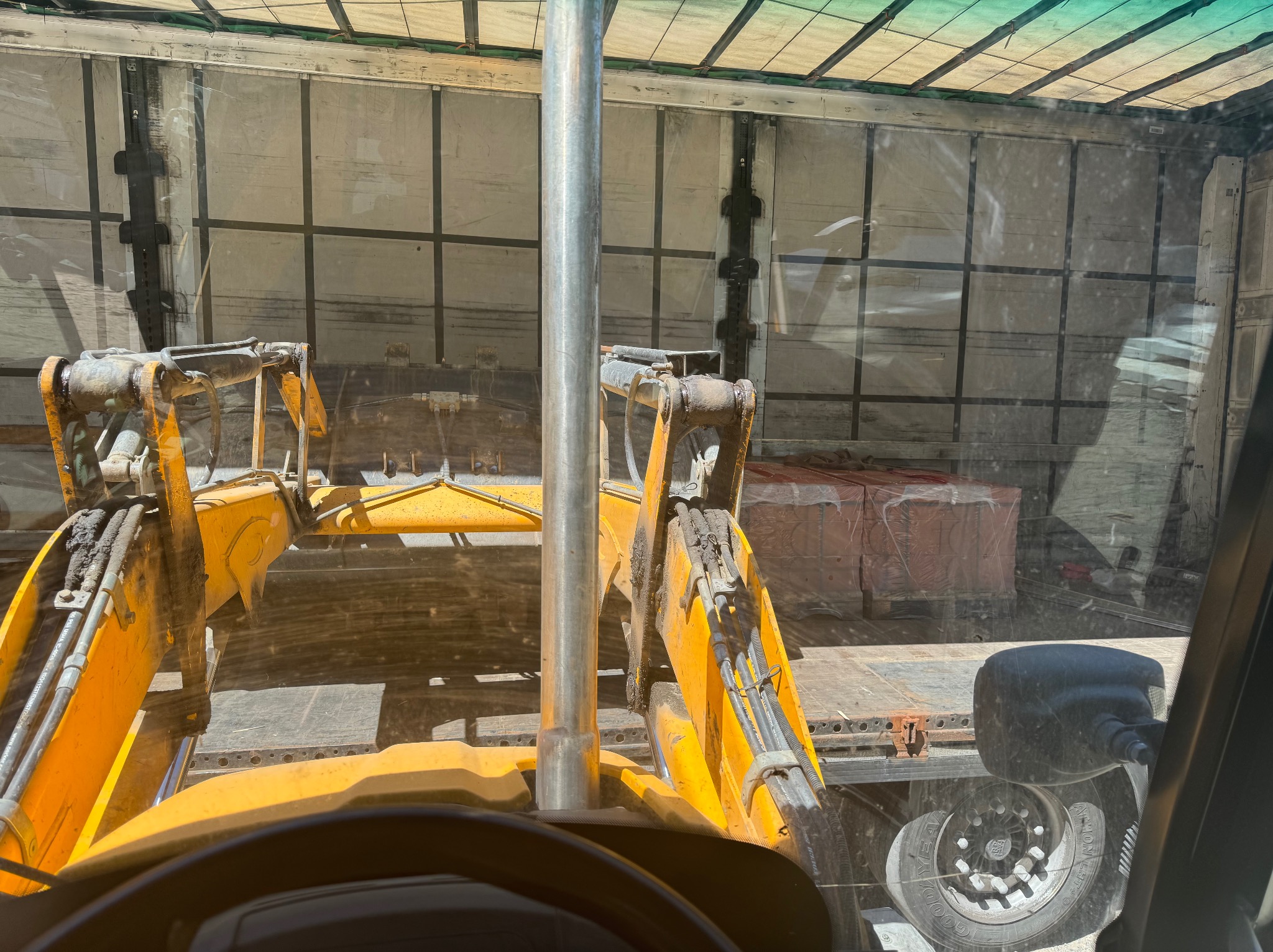 Żółta koparko-ładowarka w trakcie załadunku palet z cegłami do wnętrza naczepy ciężarówki. Widok z kabiny operatora, oświetlenie dzienne.