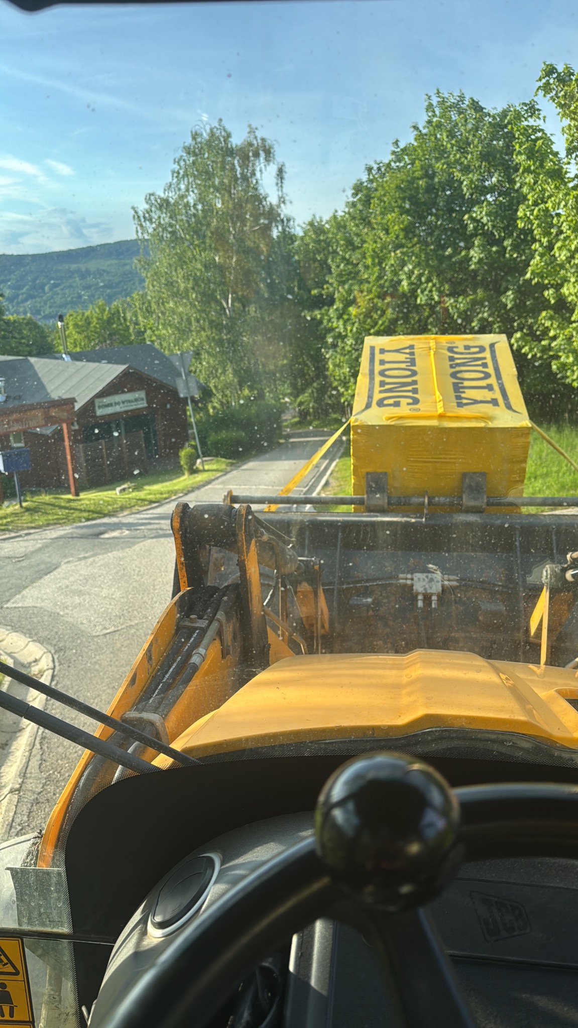 Widok z kabiny ładowarki JCB transportującej paletę bloczków Ytong na tle zieleni i zabudowań. Ujęcie z perspektywy operatora, w tle droga i budynek.