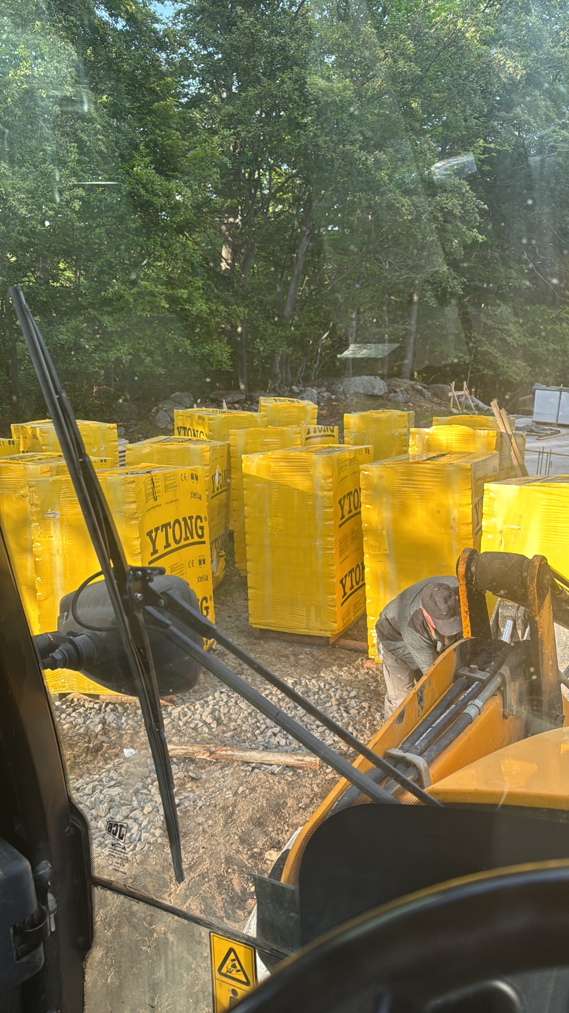 Widok z kabiny koparki na plac budowy z ułożonymi paletami bloczków Ytong, przygotowanymi do wznoszenia murów. W tle zalesiony teren.