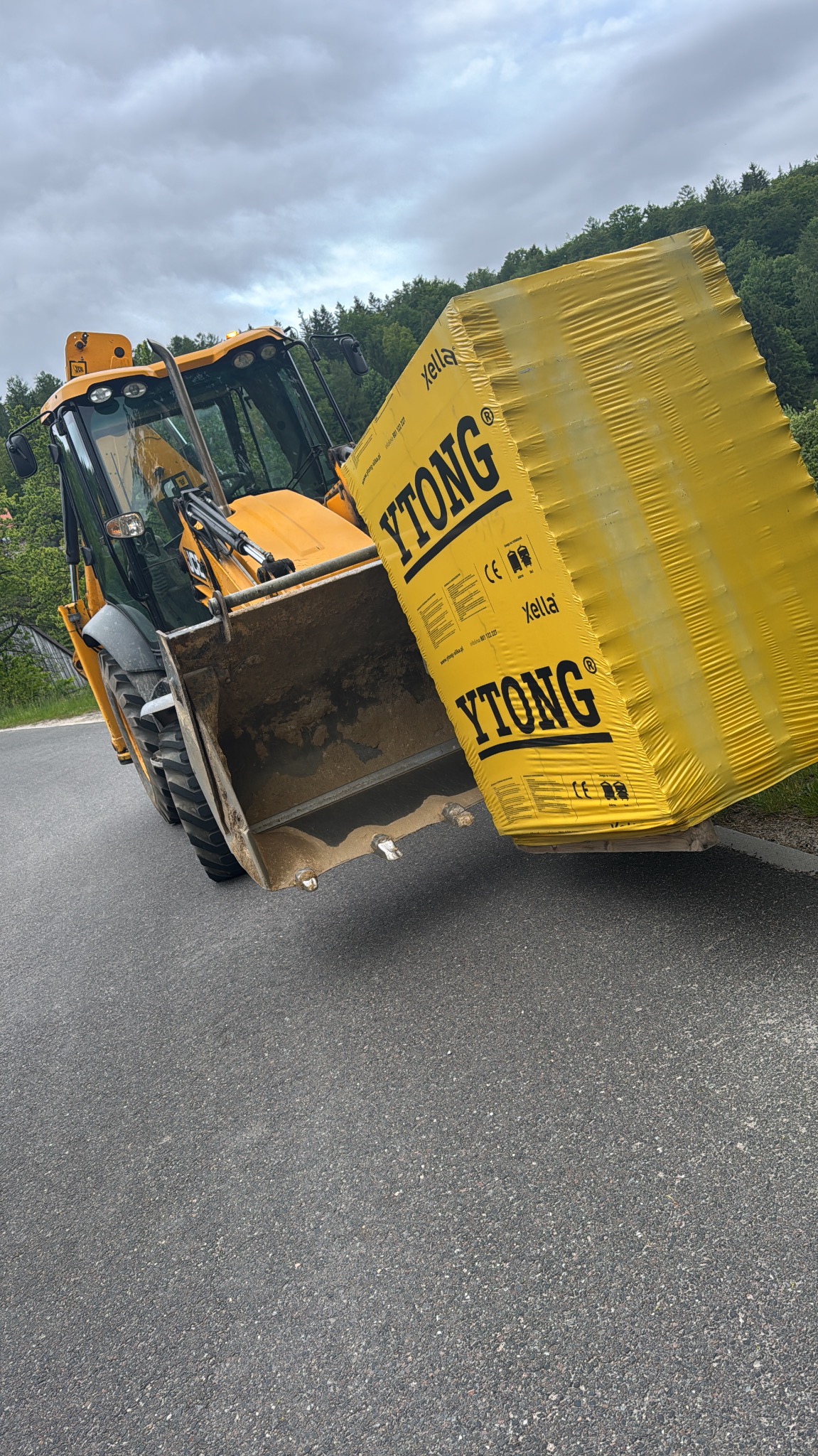 Żółta koparka z pełną łyżką bloczków Ytong na asfalcie, na tle lasu i pochmurnego nieba. Transport materiałów budowlanych.
