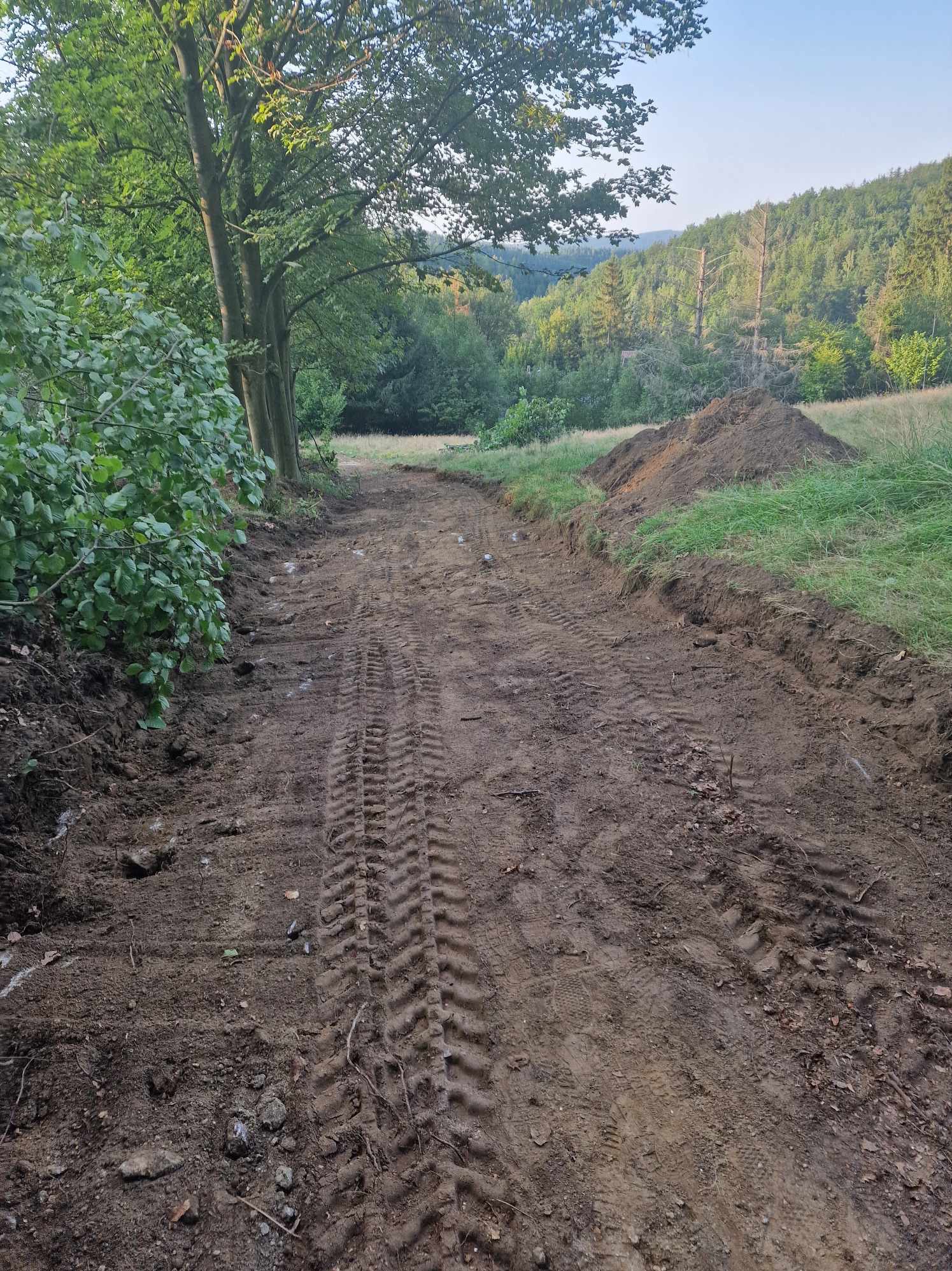 Droga gruntowa po niwelacji, widoczne ślady opon, pryzma ziemi z boku, zalesione wzgórza w tle, drzewa po lewej stronie. Krajobraz po robotach ziemnych.
