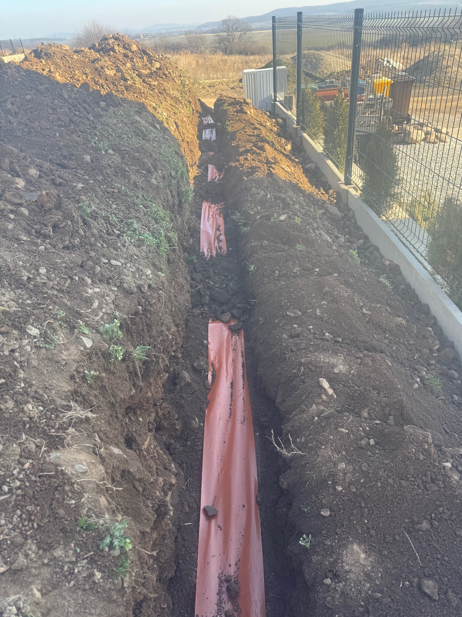 Wykopany rów z widocznymi, pomarańczowymi rurami osłonowymi. Na drugim planie ogrodzenie panelowe i niewielki budynek. Ujęcie z góry, w tle widoczne drzewa.