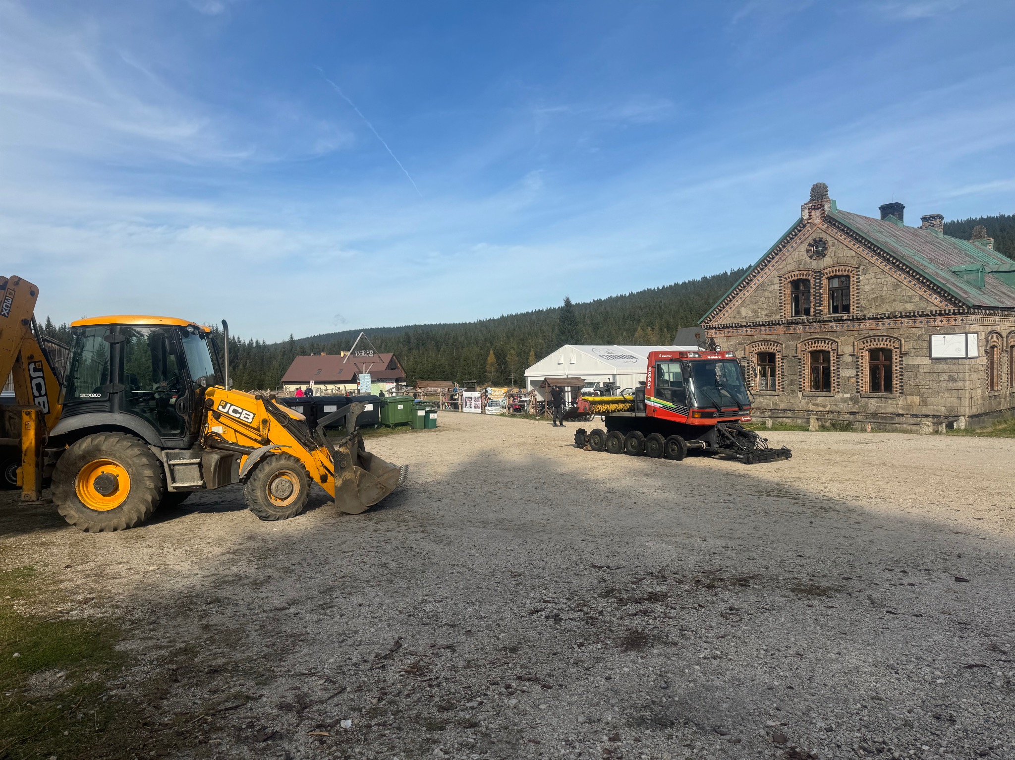 Żółta koparko-ładowarka JCB i czerwony ratrak parkują na żwirowym placu przed budynkiem z cegły i kamienia w słoneczny dzień. W tle las i błękitne niebo.