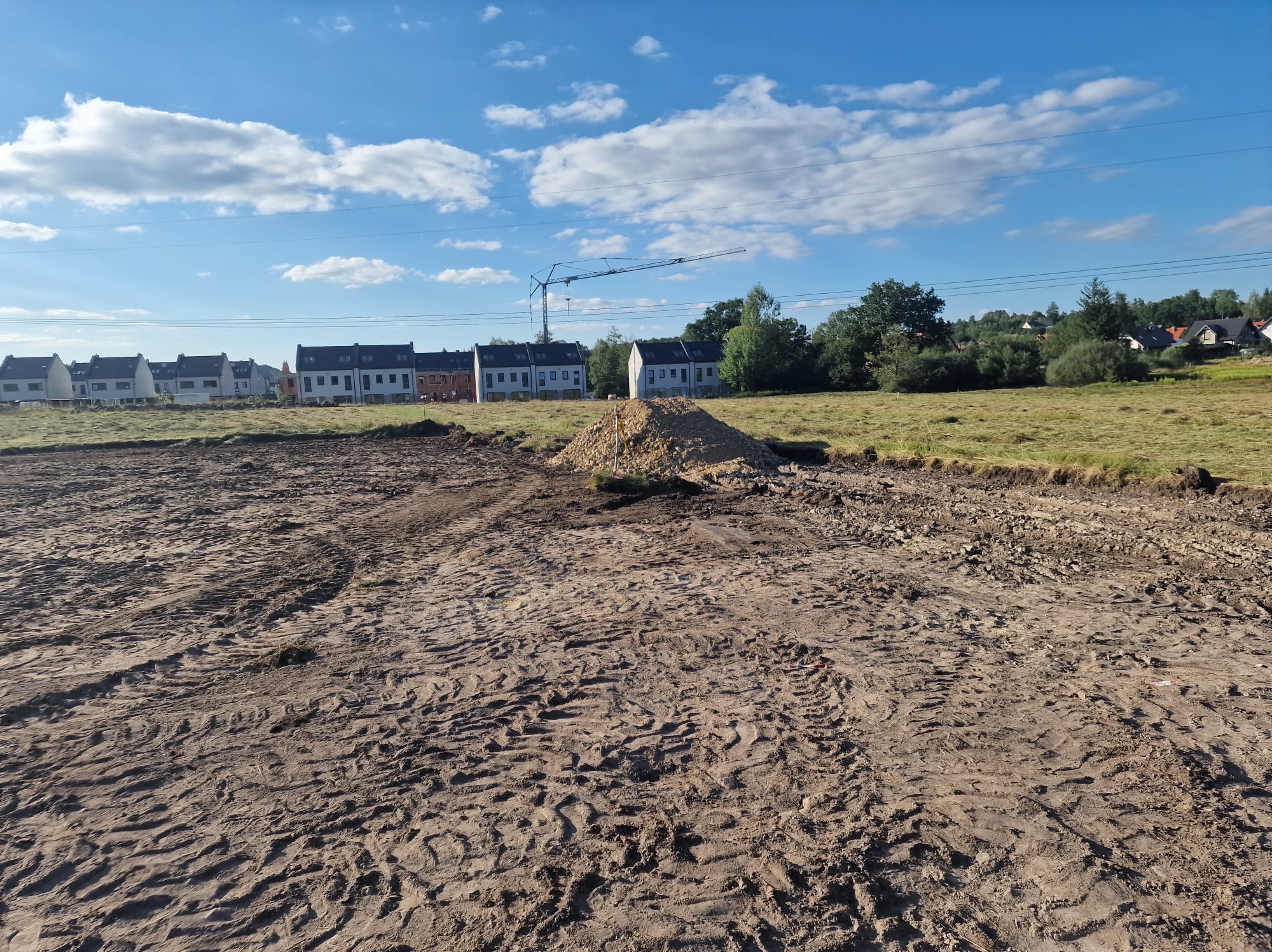 Przygotowany teren pod budowę z widocznymi śladami opon, stertą ziemi na środku i nowymi domami w tle. Na horyzoncie widoczny żuraw budowlany.