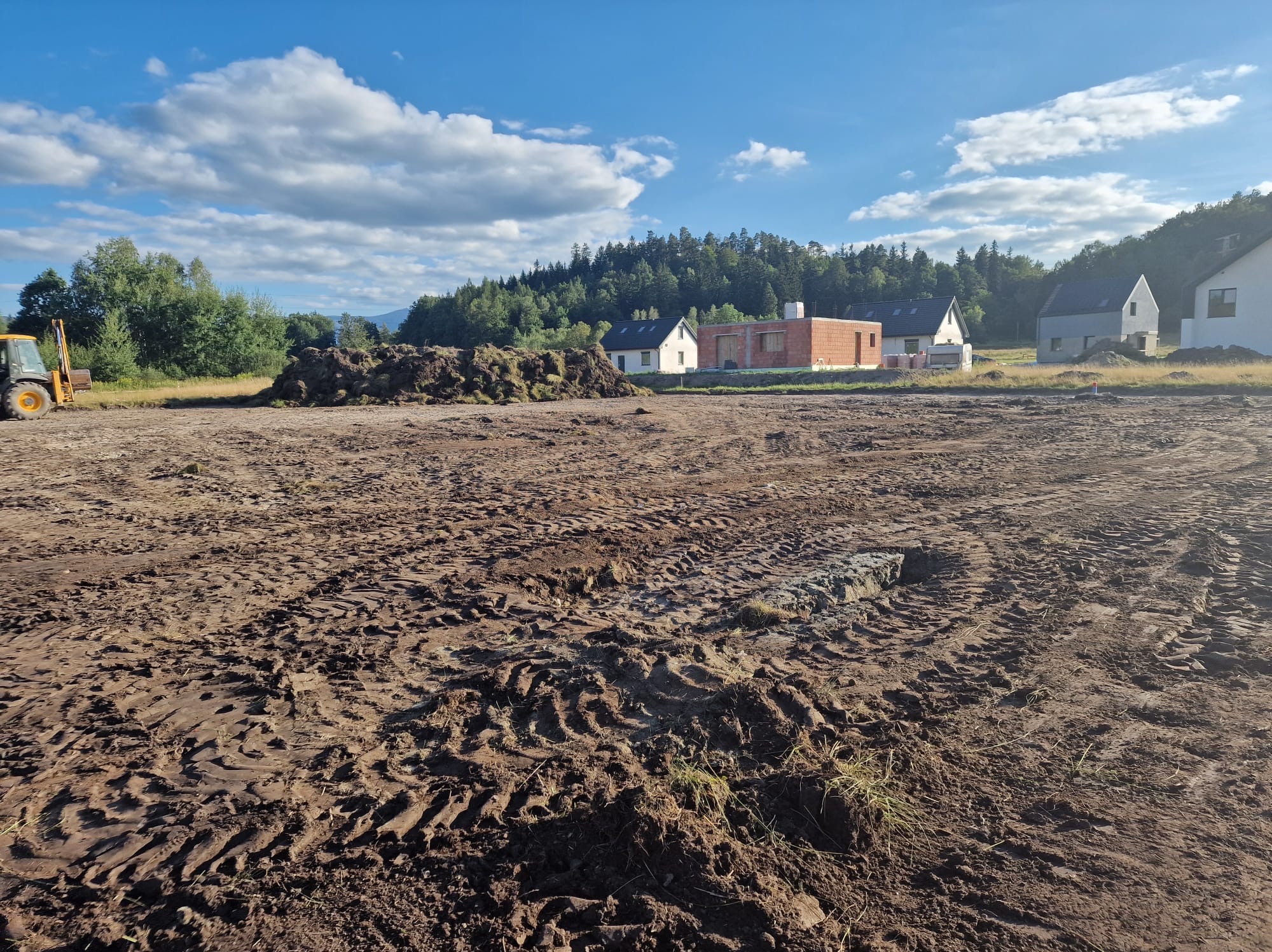 Przygotowany teren pod budowę z widocznymi śladami opon, stertą ziemi i częściowo wybudowanym budynkiem w tle. W oddali las i błękitne niebo.
