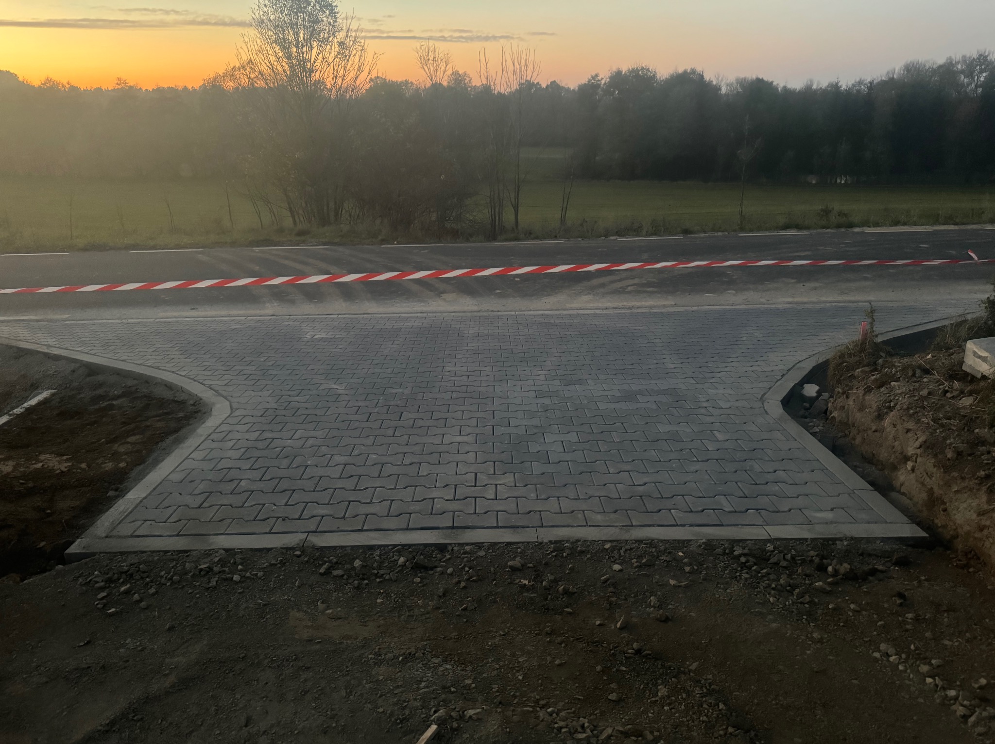 Ułożona szara kostka brukowa na rozwidleniu drogi, zabezpieczona taśmą ostrzegawczą; w tle drzewa i niebo o zachodzie słońca. Widoczne nierówności terenu po bokach.