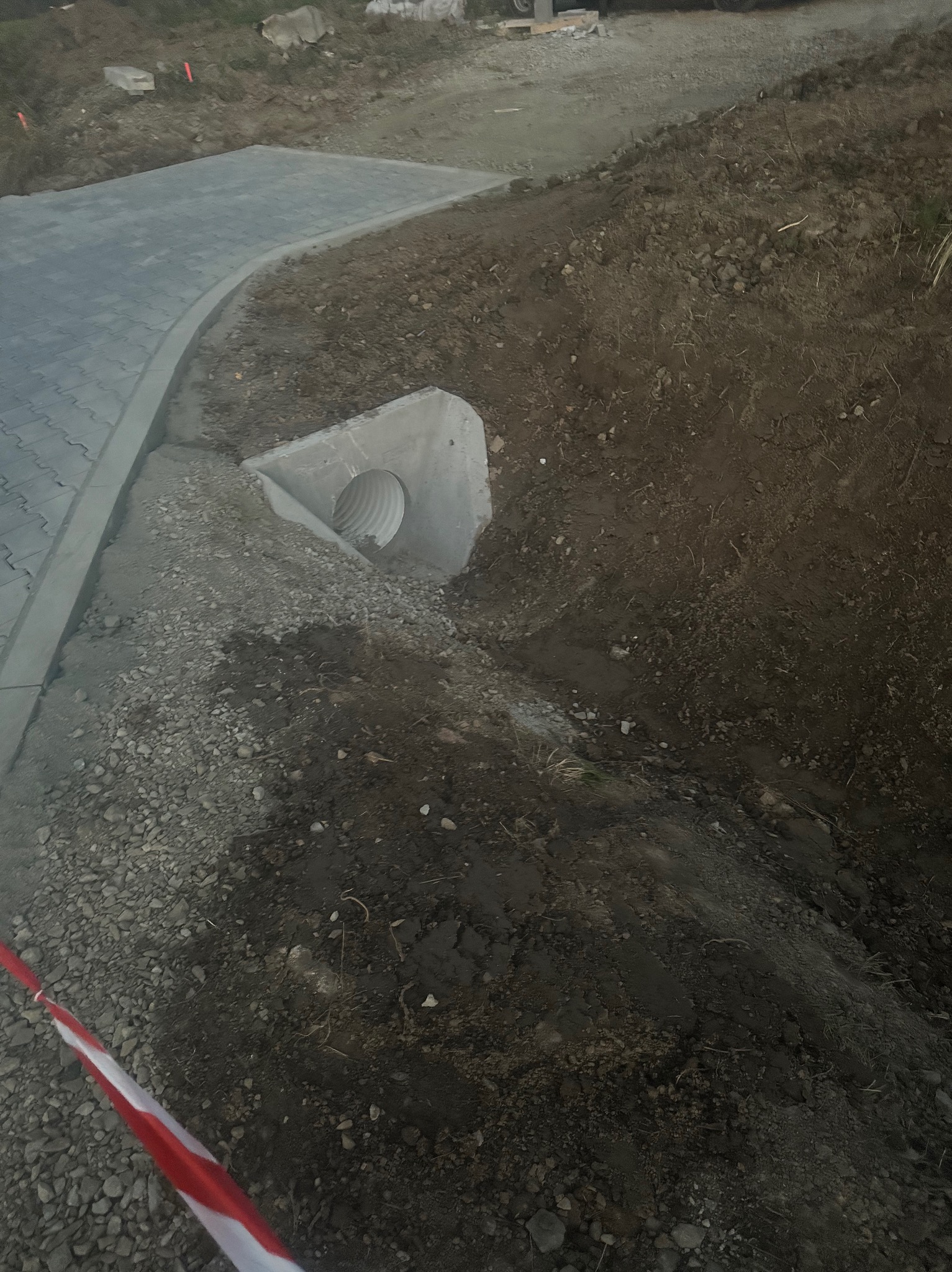 Ułożona kostka brukowa z betonowym korytkiem odpływowym. Wykop ziemny z widoczną rurą drenażową. Taśma ostrzegawcza w rogu kadru.