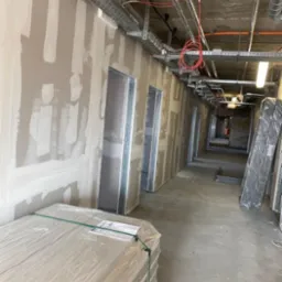 Korytarz w trakcie wykańczania, ściany pokryte szpachlą gipsową, widoczne metalowe profile ościeżnic, instalacje pod sufitem, paleta płyt gipsowo-kartonowych w folii na pierwszym planie.