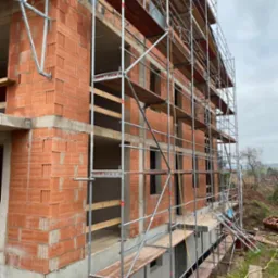 Surowy stan budynku dwukondygnacyjnego z czerwonej cegły, otoczonego rusztowaniami, widok z zewnątrz na tle pochmurnego nieba i zieleni.