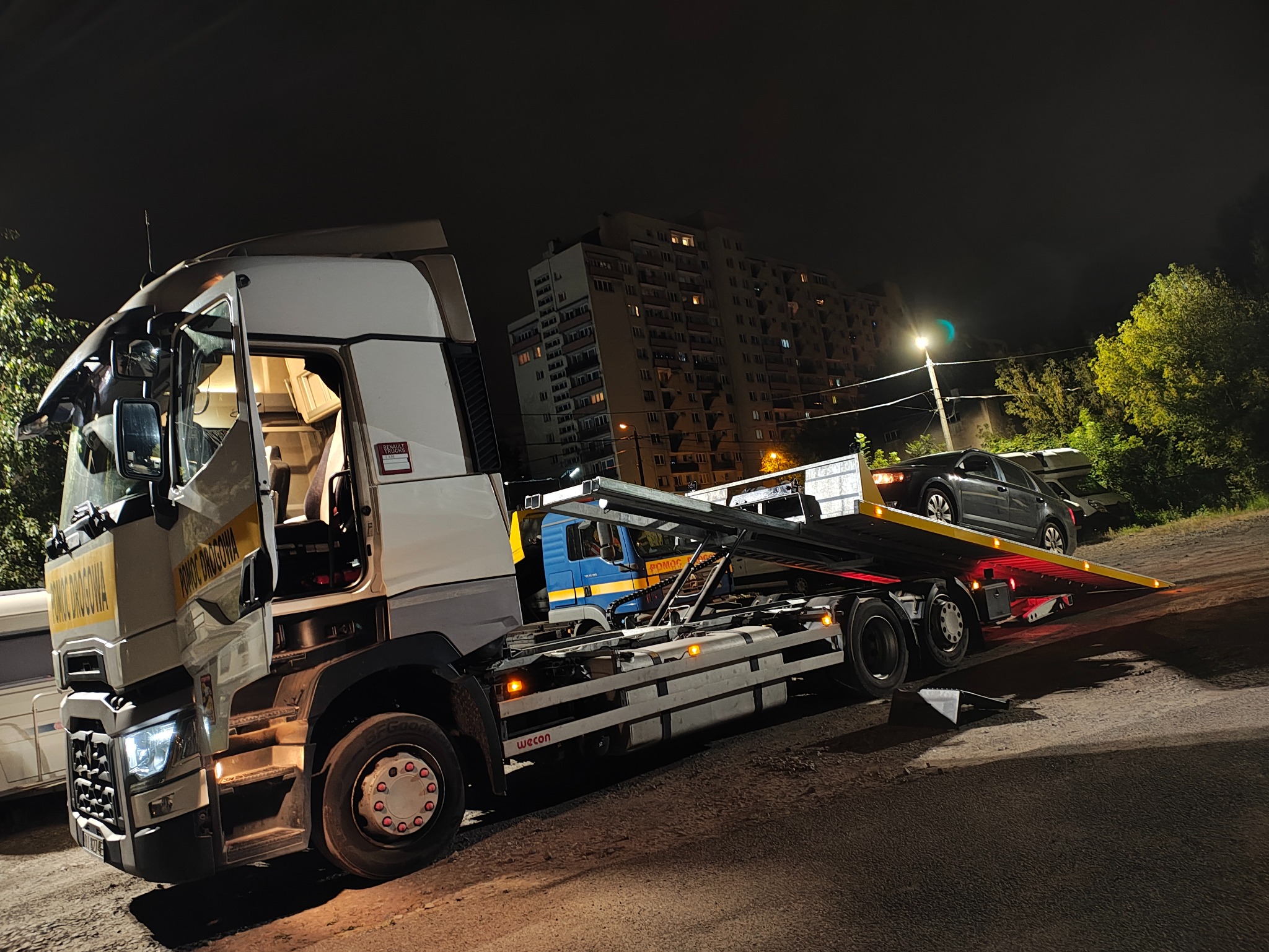 Nocna scena z lawetą Renault Trucks z otwartymi drzwiami kabiny, transportującą szary samochód osobowy na tle bloku mieszkalnego i drzew.