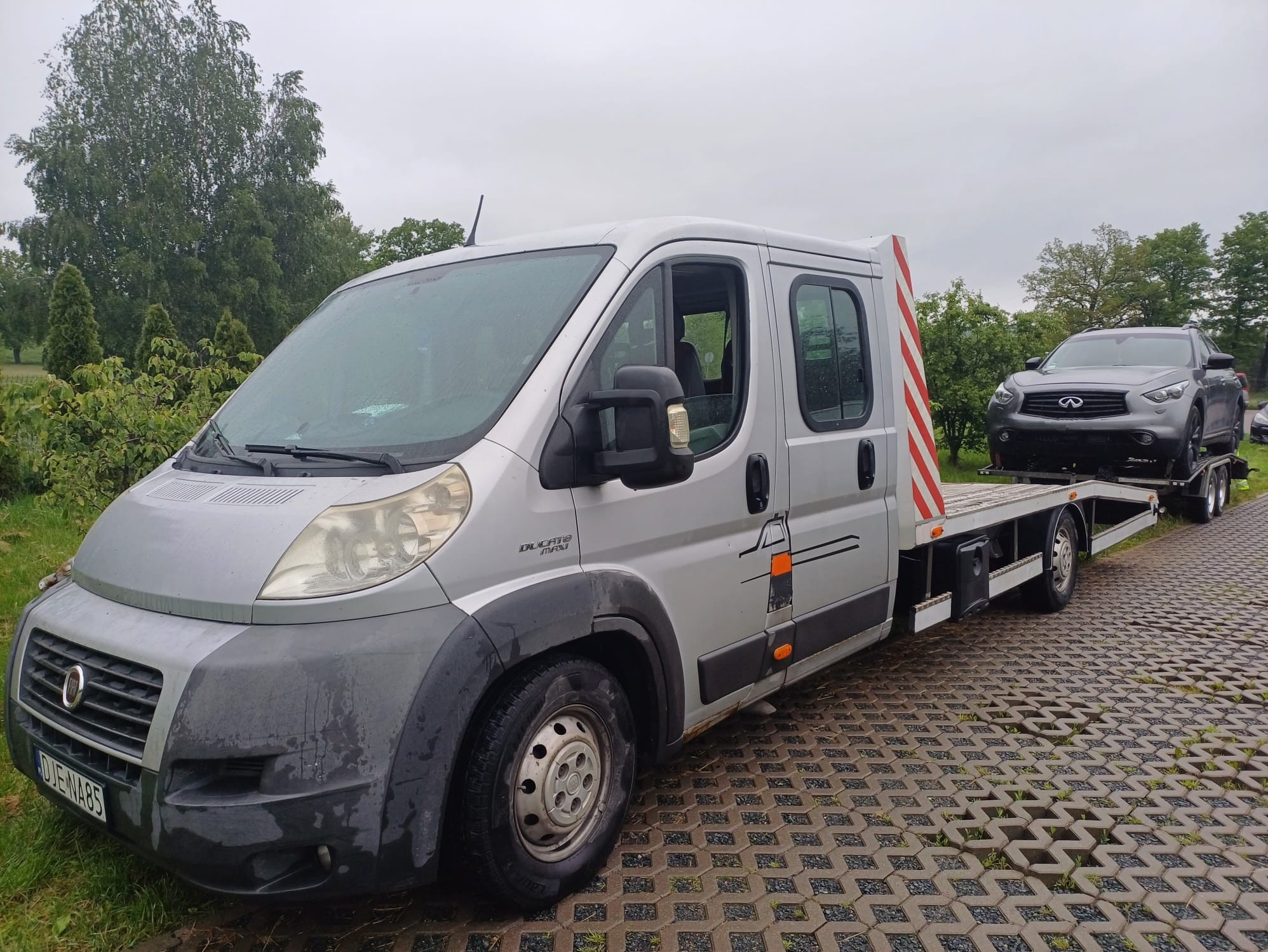 Srebrna laweta Fiat Ducato z zaparkowanym szarym Infiniti na platformie, stojąca na betonowej kostce w deszczowy dzień. Widoczne drzewa w tle.