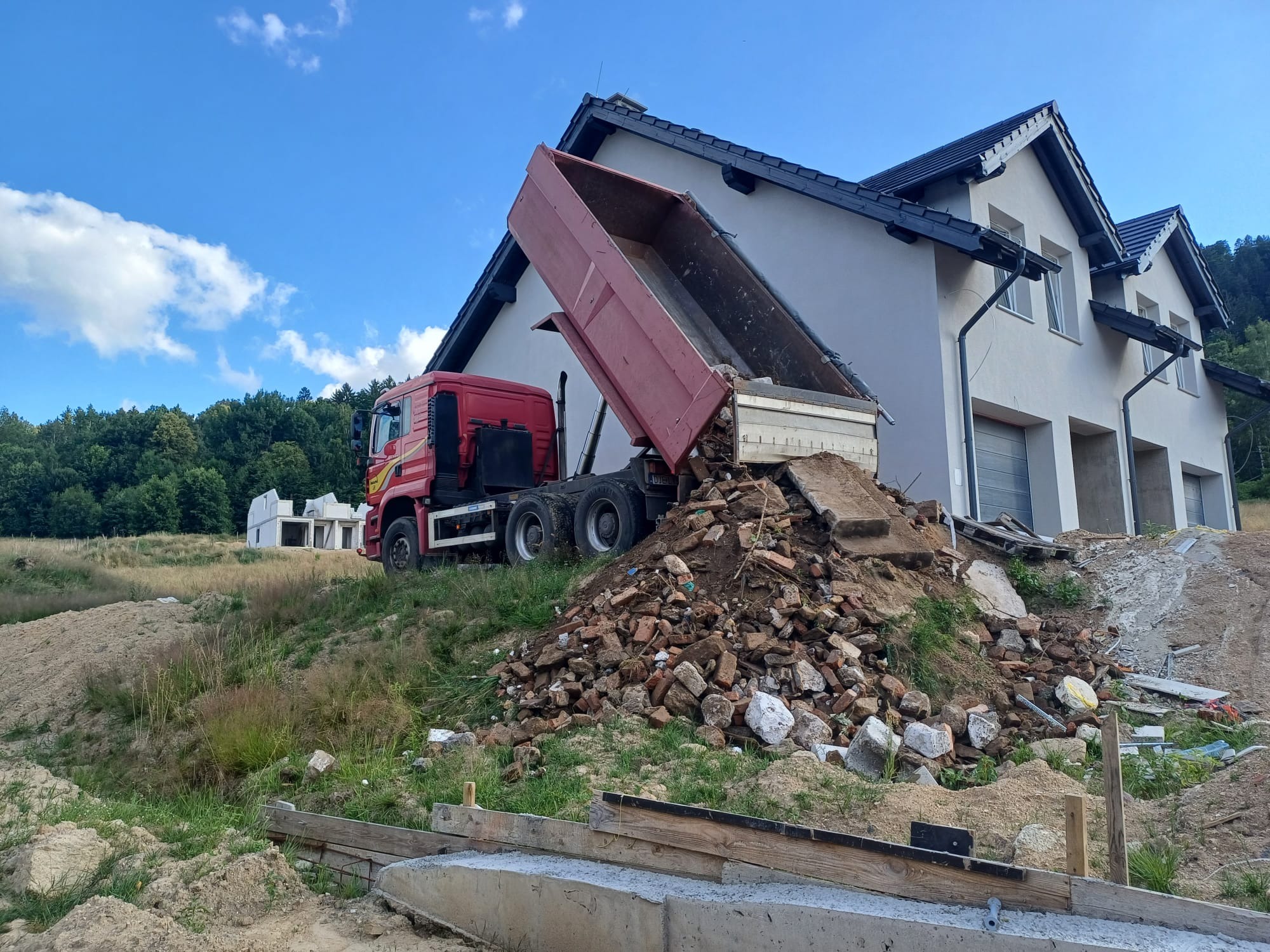 Czerwona wywrotka z podniesioną skrzynią wysypuje gruz na tle budowanego domu jednorodzinnego, widok z boku w słoneczny dzień.