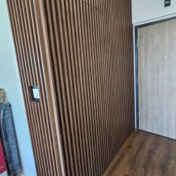 POCO Meble Patrycja Bubiłek - Drewniana ściana z lameli w przedpokoju, z ukrytymi gniazdkami i włącznikiem, na tle drzwi wejściowych i podłogi z paneli. Nowoczesny design.