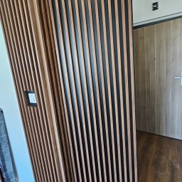 POCO Meble Patrycja Bubiłek - Drewniane lamele na czarnym tle tworzą nowoczesną ściankę działową. Widoczne gniazdko elektryczne i fragment drzwi. Podłoga z paneli imitujących drewno.