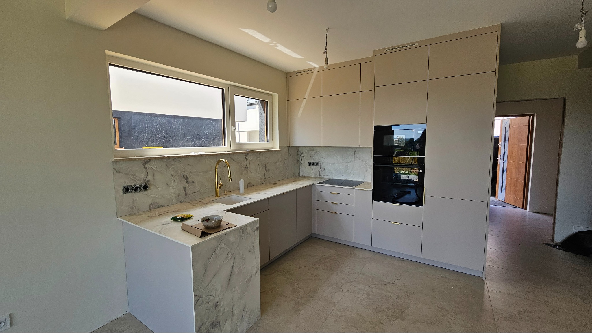 Jasna kuchnia na wymiar z marmurowym blatem i złotą armaturą. Widoczne okno z widokiem na zewnątrz i sprzęt AGD w zabudowie. Minimalistyczny design z elementami luksusu.