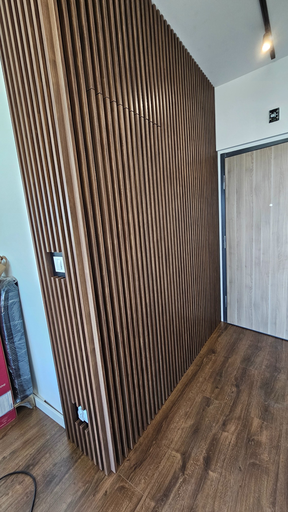 Drewniana ściana z lameli w przedpokoju, z ukrytymi gniazdkami i włącznikiem, na tle drzwi wejściowych i podłogi z paneli. Nowoczesny design.