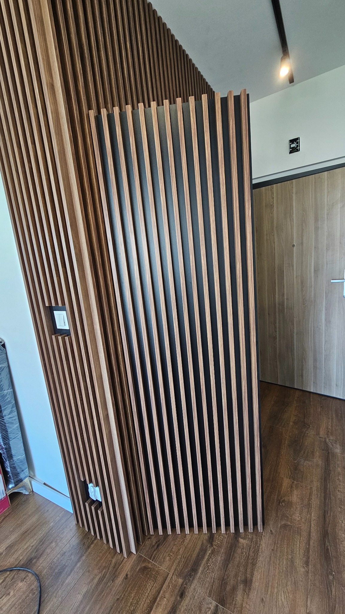 Drewniane lamele na czarnym tle tworzą nowoczesną ściankę działową. Widoczne gniazdko elektryczne i fragment drzwi. Podłoga z paneli imitujących drewno.