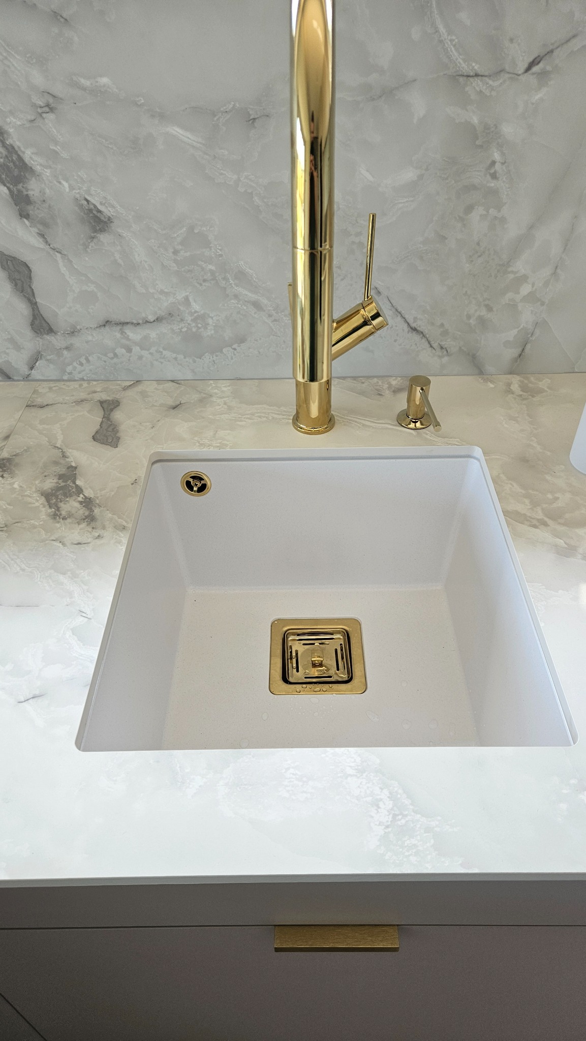 Biały, kwadratowy zlew granitowy z złotą armaturą i dozownikiem mydła na blacie z marmurowym wzorem. U dołu widoczny fragment szarej szafki z złotym uchwytem.