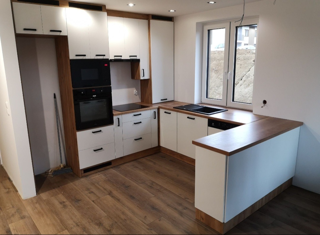 Nowoczesna kuchnia z białymi frontami i drewnianymi akcentami. Widoczne zabudowane sprzęty AGD, czarne uchwyty i blat z ciemnego drewna. Podłoga z paneli imitujących drewno.