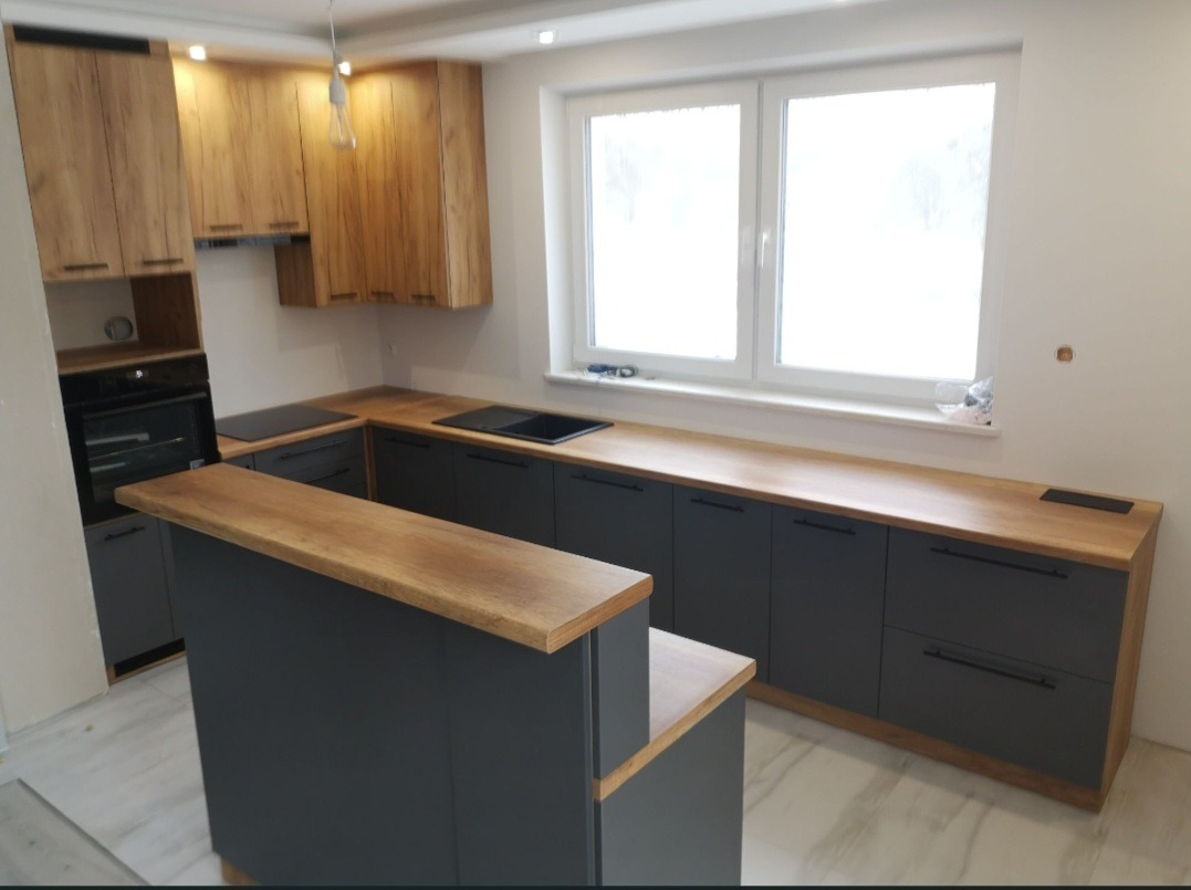 Nowoczesna kuchnia z szarymi szafkami i drewnianym blatem, oświetlona wiszącą lampą. Widok na okno z białymi ramami i marmurową podłogę. Minimalistyczny design.