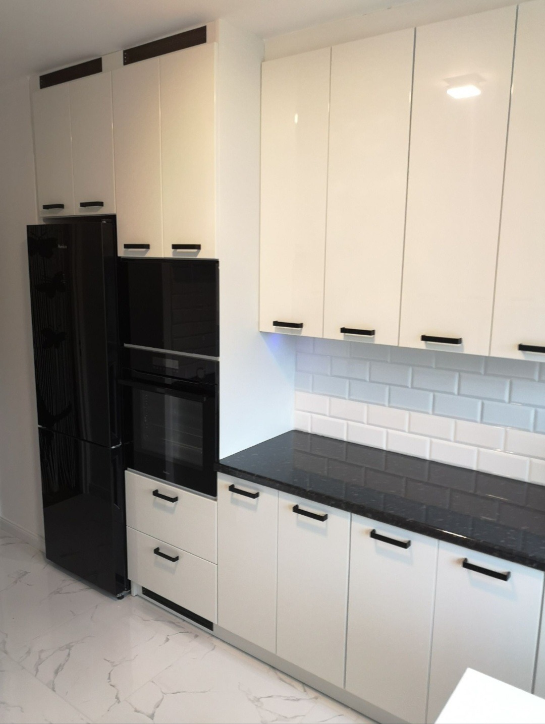 Biała, nowoczesna zabudowa kuchenna z czarnym blatem i sprzętem AGD. Minimalistyczne uchwyty, płytki typu metro. Widok na marmurową podłogę.