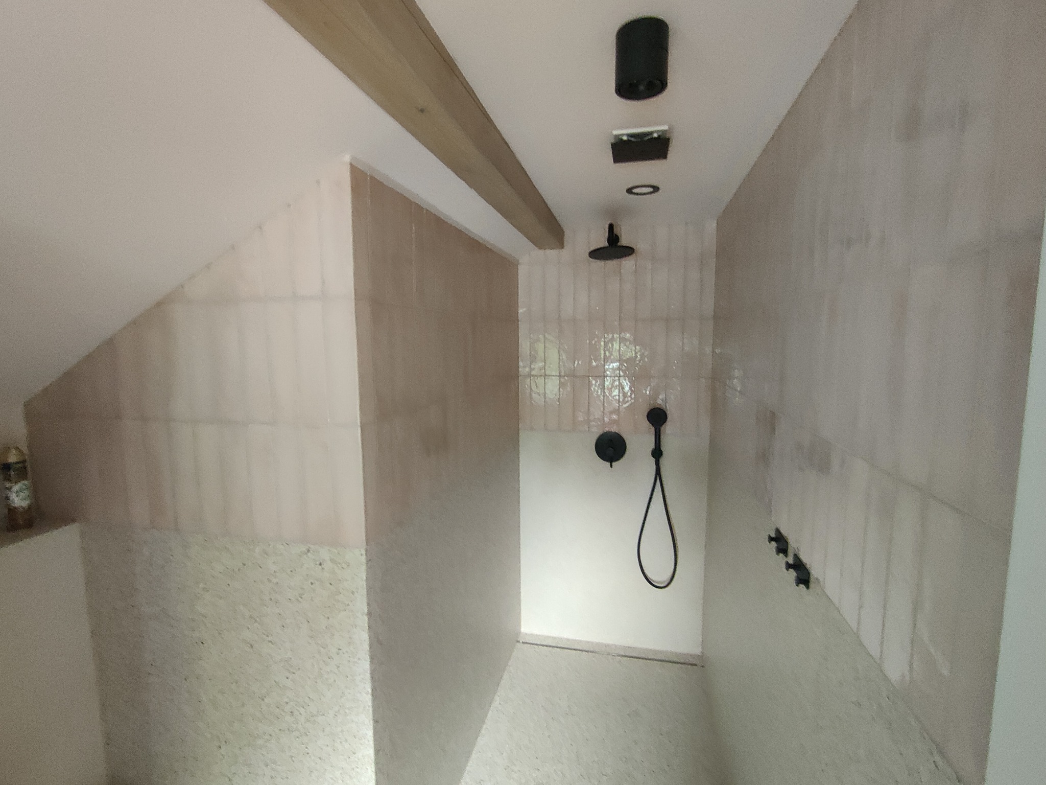 Wykończona kabina prysznicowa z beżowymi, podłużnymi płytkami na ścianach i mozaiką na dole, czarna armatura, drewniana belka stropowa i skośny sufit.