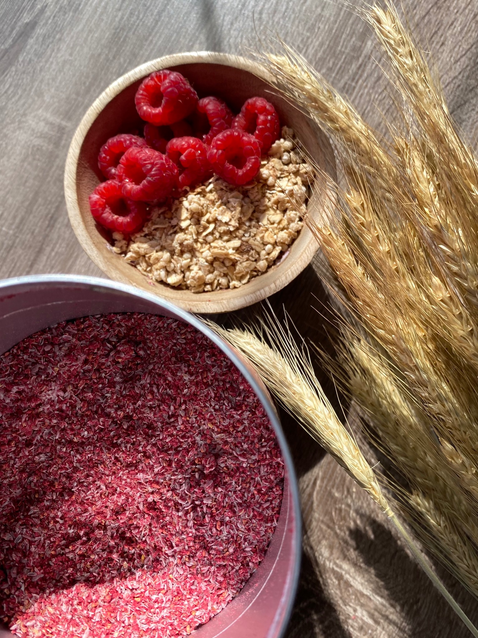 Fotografia produktowa: owsianka z malinami w drewnianej miseczce, czerwony błonnik w puszce i kłosy pszenicy na drewnianym stole w ciepłym świetle.