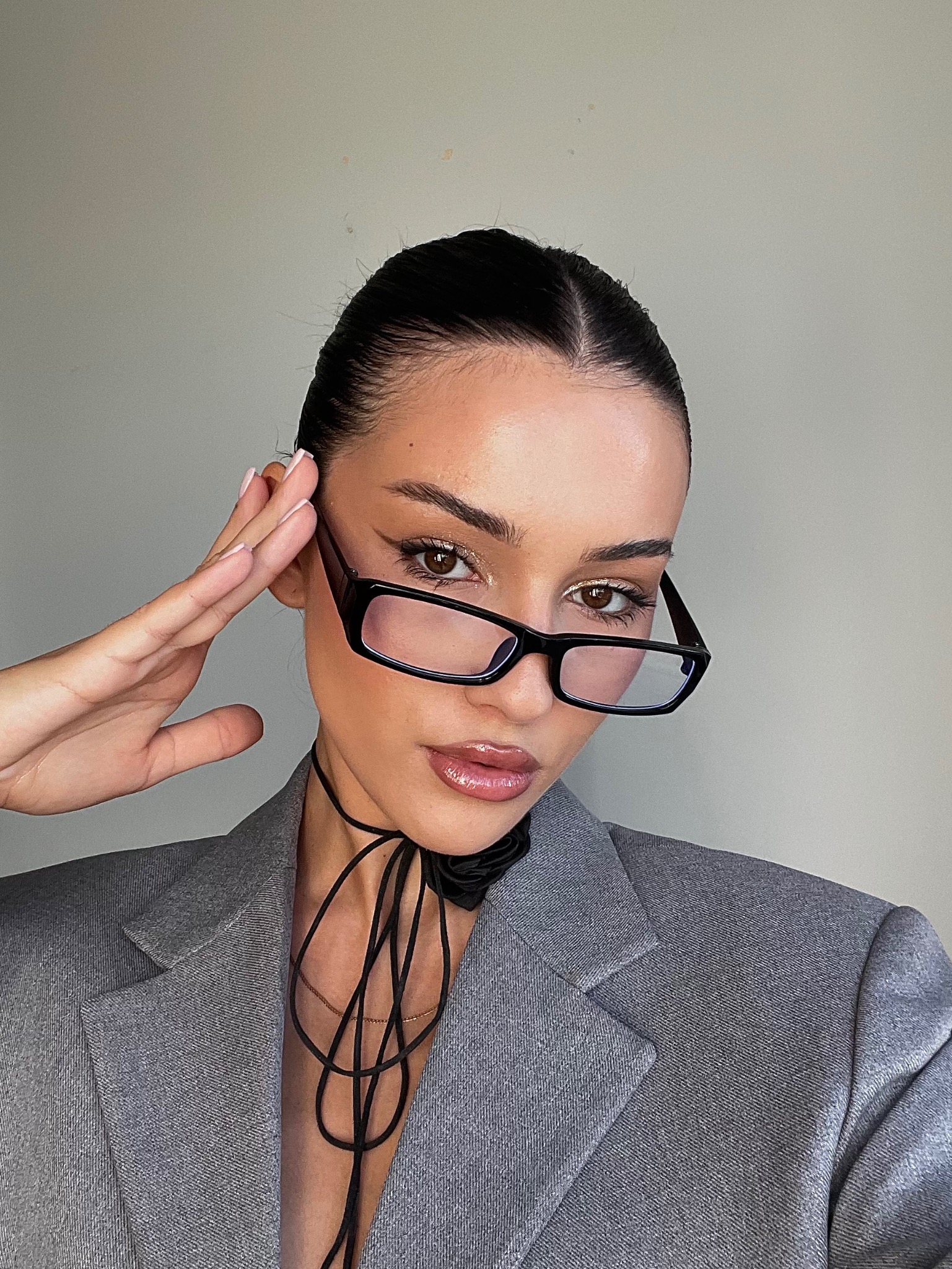 Portret kobiety w okularach i szarym żakiecie prezentujący makijaż, z wyraźnie podkreślonym okiem i błyszczącymi ustami, na neutralnym tle.