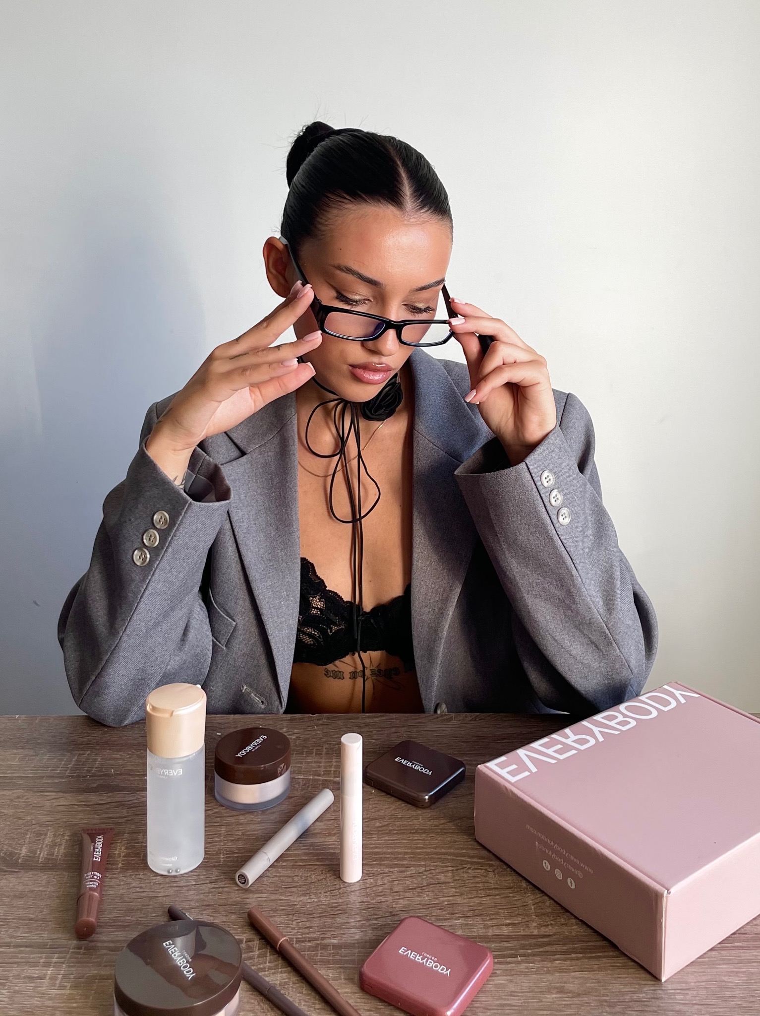 Sesja produktowa kosmetyków EverybodyLondon: modelka w marynarce i okularach prezentuje zestaw kosmetyków na drewnianym stole, z pudełkiem firmowym w tle.