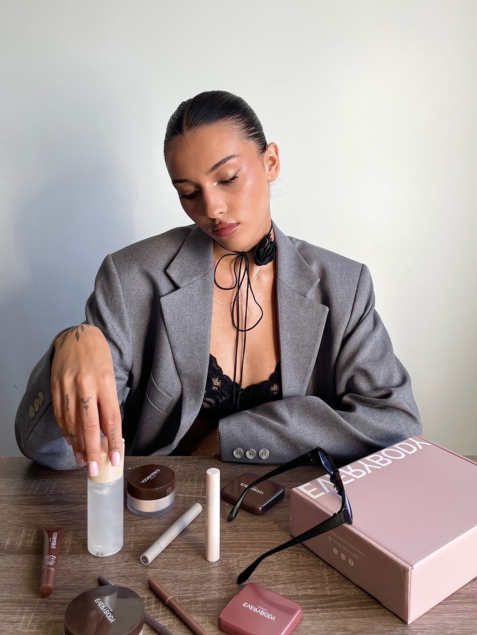 Sesja produktowa kosmetyków EverybodyLondon: modelka w szarym żakiecie prezentuje nową kolekcję na drewnianym stole, z pudrowym pudełkiem i okularami obok.