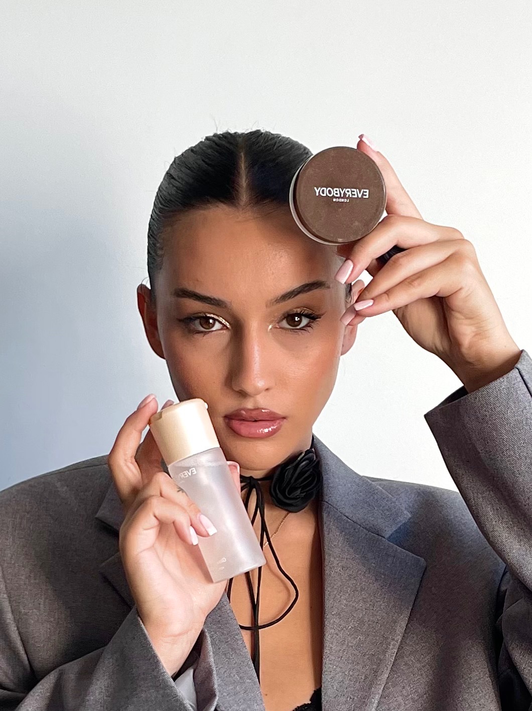 Portret kobiety w marynarce, prezentującej kosmetyki Everybody London: puder w dłoni nad głową i flakon z atomizerem w drugiej dłoni. Uwagę zwraca makijaż.